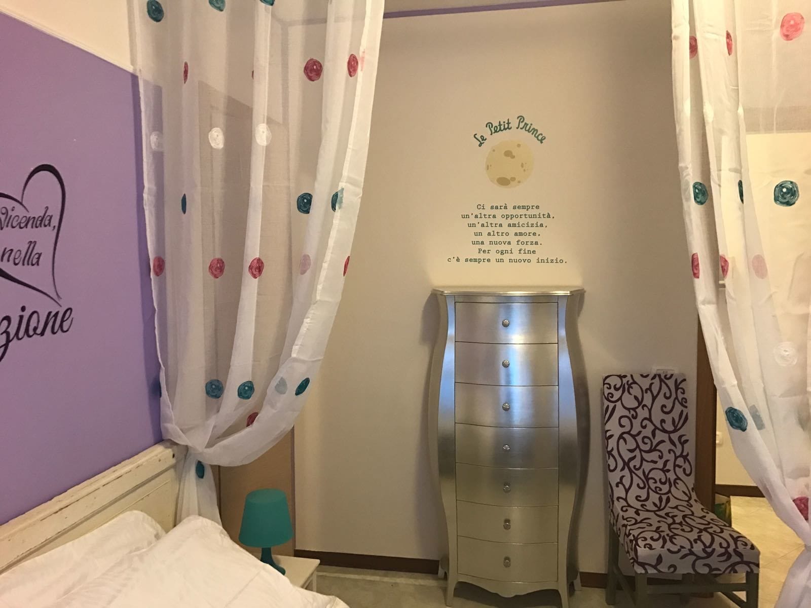 Un dormitorio con pared morada, una cama con ropa blanca, un tocador, una silla y una pared con una luna y texto.