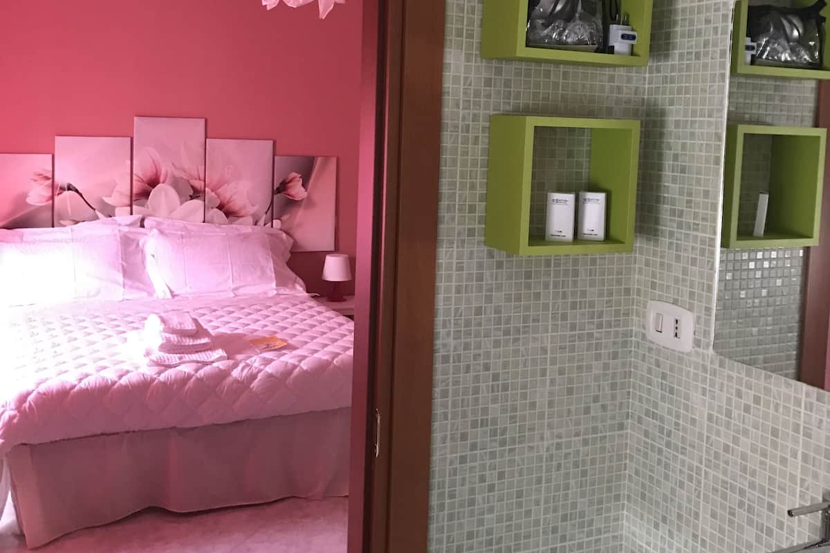 A bedroom with a pink bedspread, a bedside table with a lamp, and a pink flower decoration. There is a bathroom with a glass sink, a mirror, and green storage shelves.