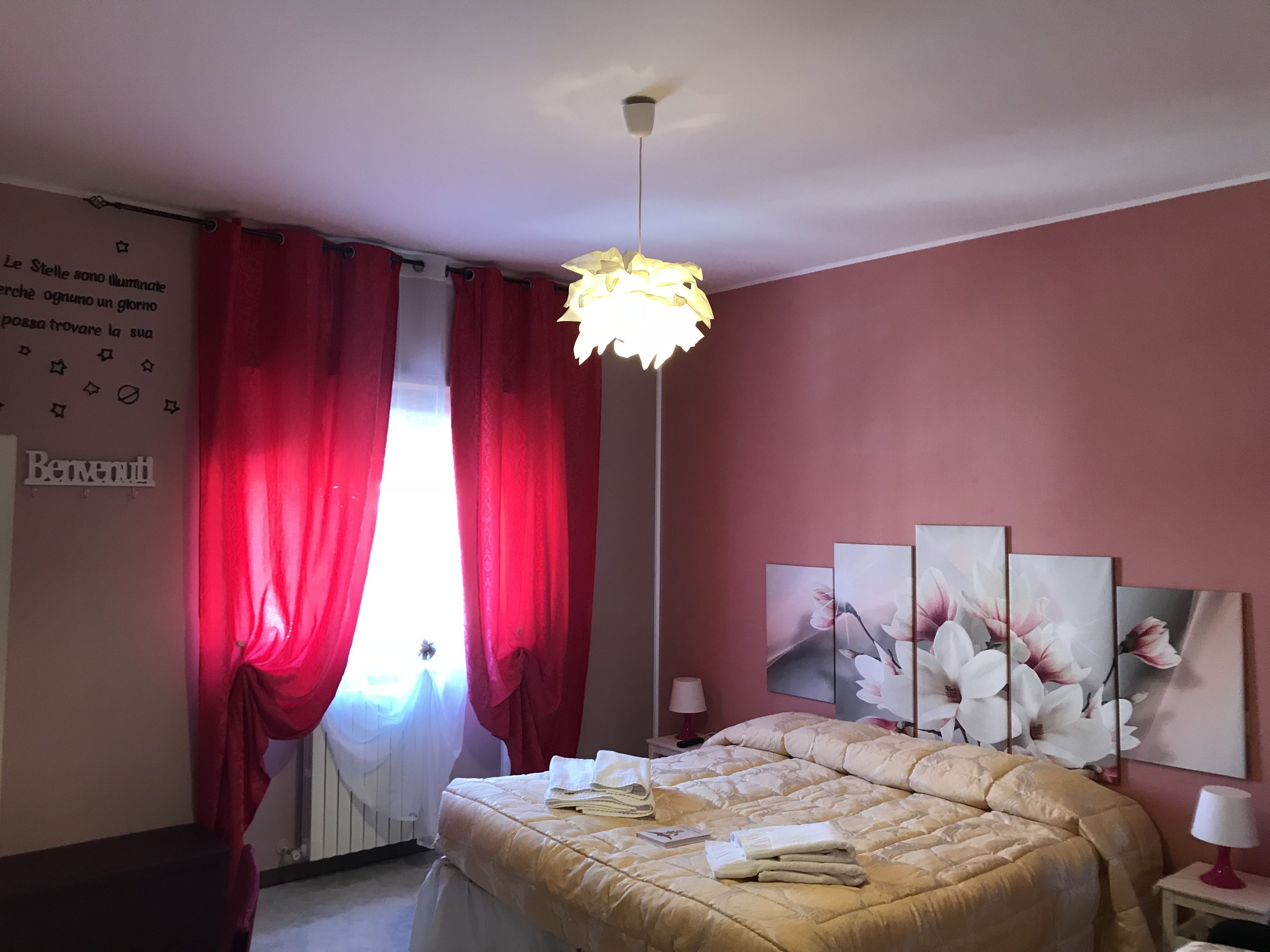 A bedroom with a bed, red curtains, a wall-mounted light fixture, and floral artwork.