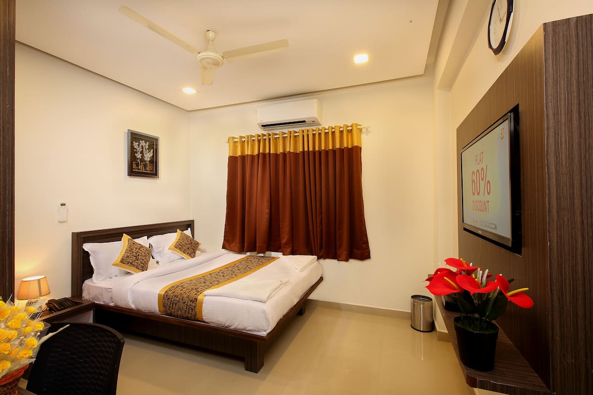 A hotel room with a bed, a desk, a chair, a plant, a clock, and an air conditioning unit.