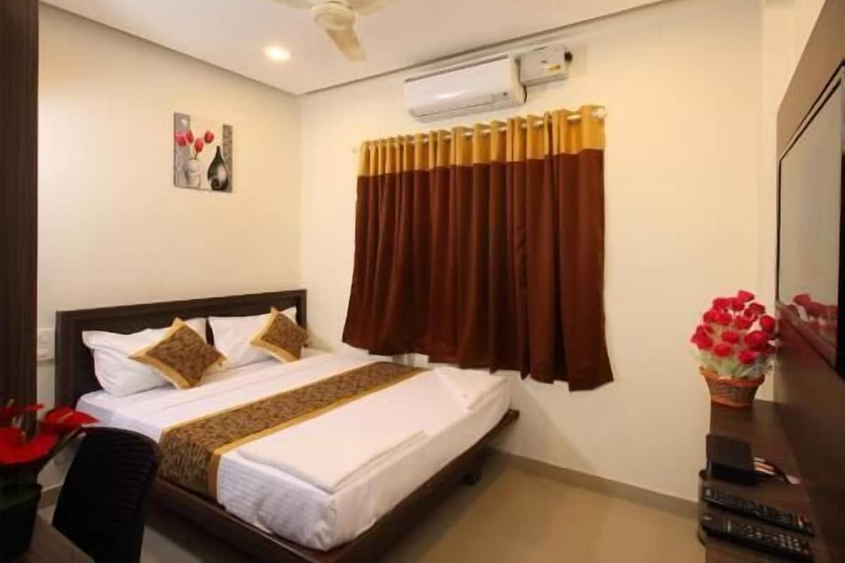 A hotel room with a bed, a ceiling fan, an air conditioning unit, a window with curtains, a wall-mounted TV, and a small table with a vase of flowers.