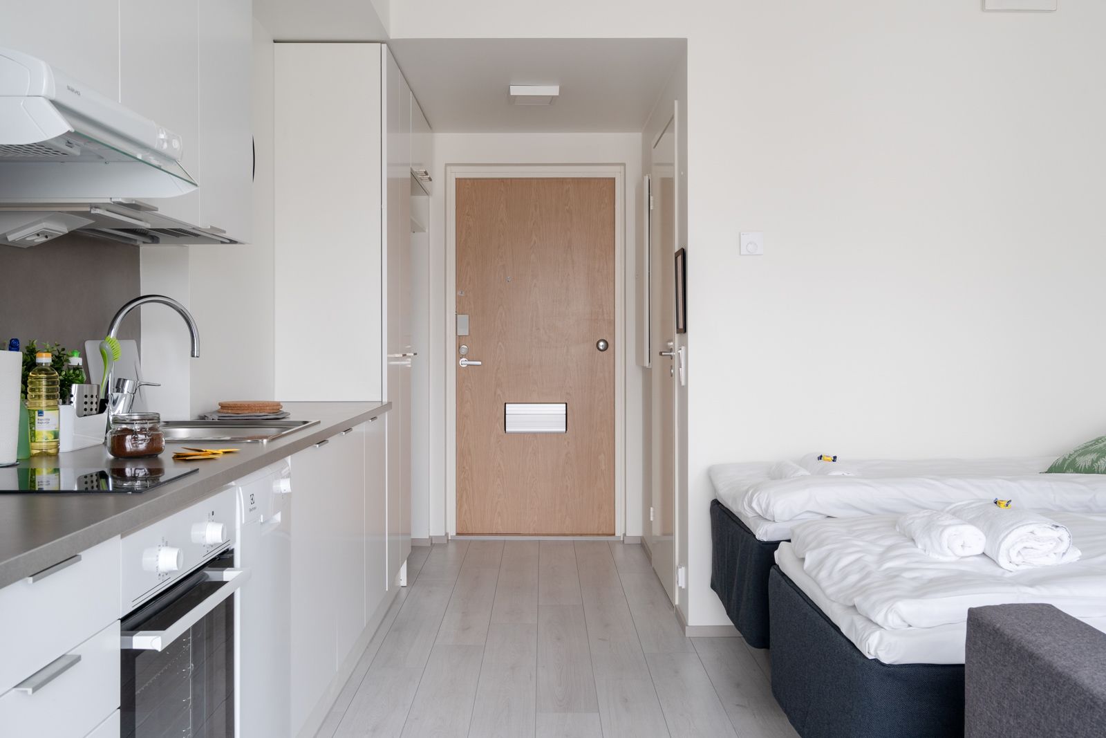A compact hotel room with a kitchenette, a bed with neatly folded white linens, and a wooden door.
