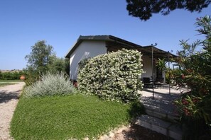 A house with a white exterior, a covered patio with outdoor furniture, and a well-maintained garden with various shrubs and a lawn. - Agriturismo Peretti (Orbetello)