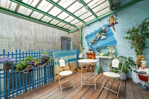A covered patio area with a blue fence, two white metal chairs, a small round table, and various potted plants.