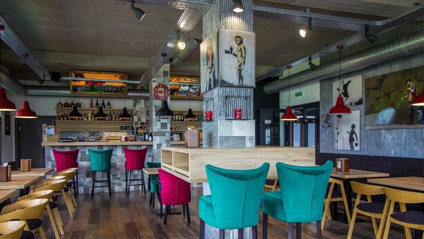A modern restaurant interior with wooden tables and colorful chairs, a bar area with bottles, and a mural on the wall. - Hotel & Bistro 63 (Poprad)