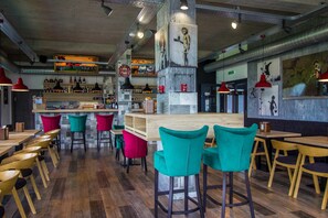 Un restaurante moderno con mesas de madera y sillas coloridas, una zona de bar con botellas y un mural en la pared.