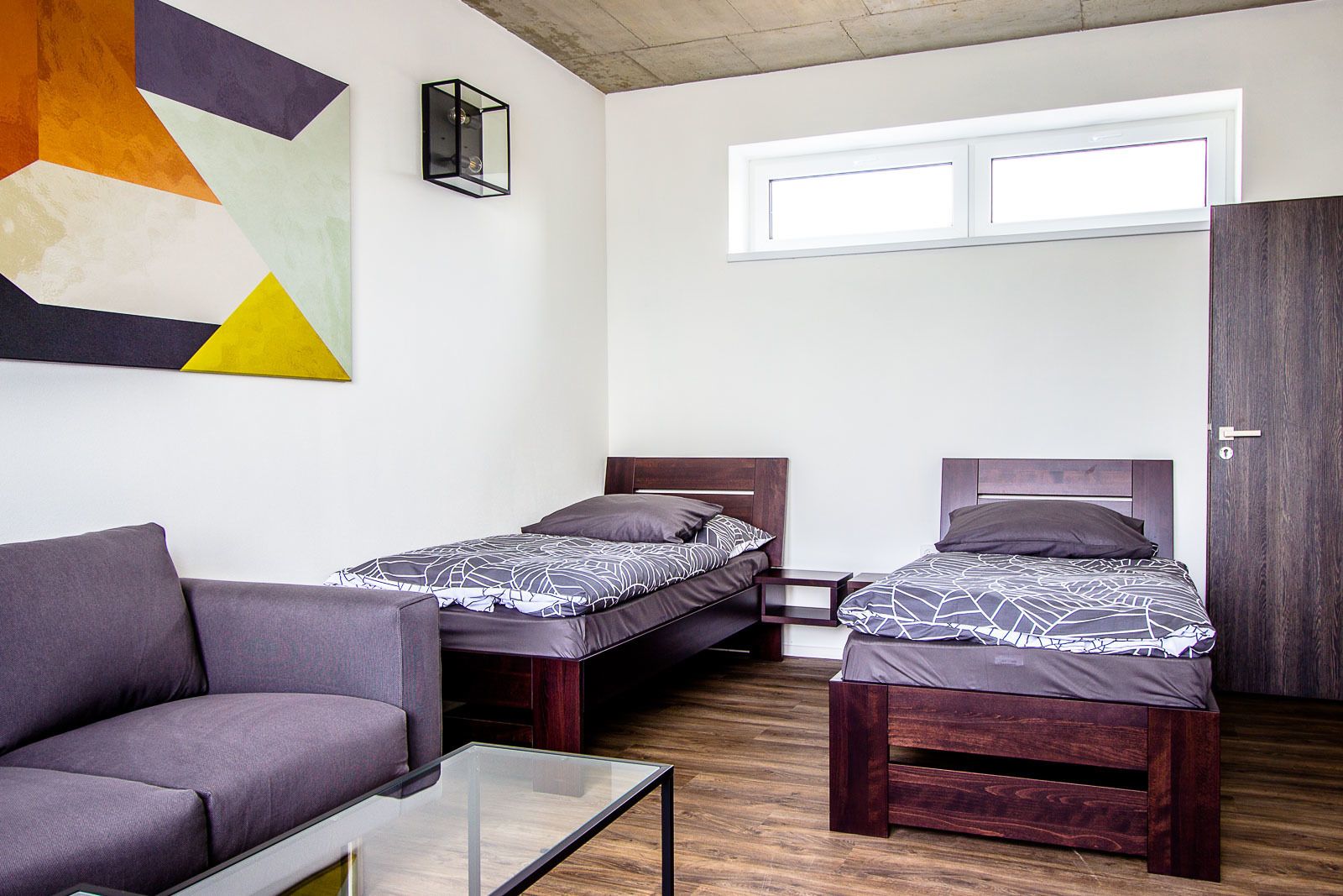 Habitación compacta con dos camas individuales, un sofá, una mesita y una obra de arte geométrica en la pared.