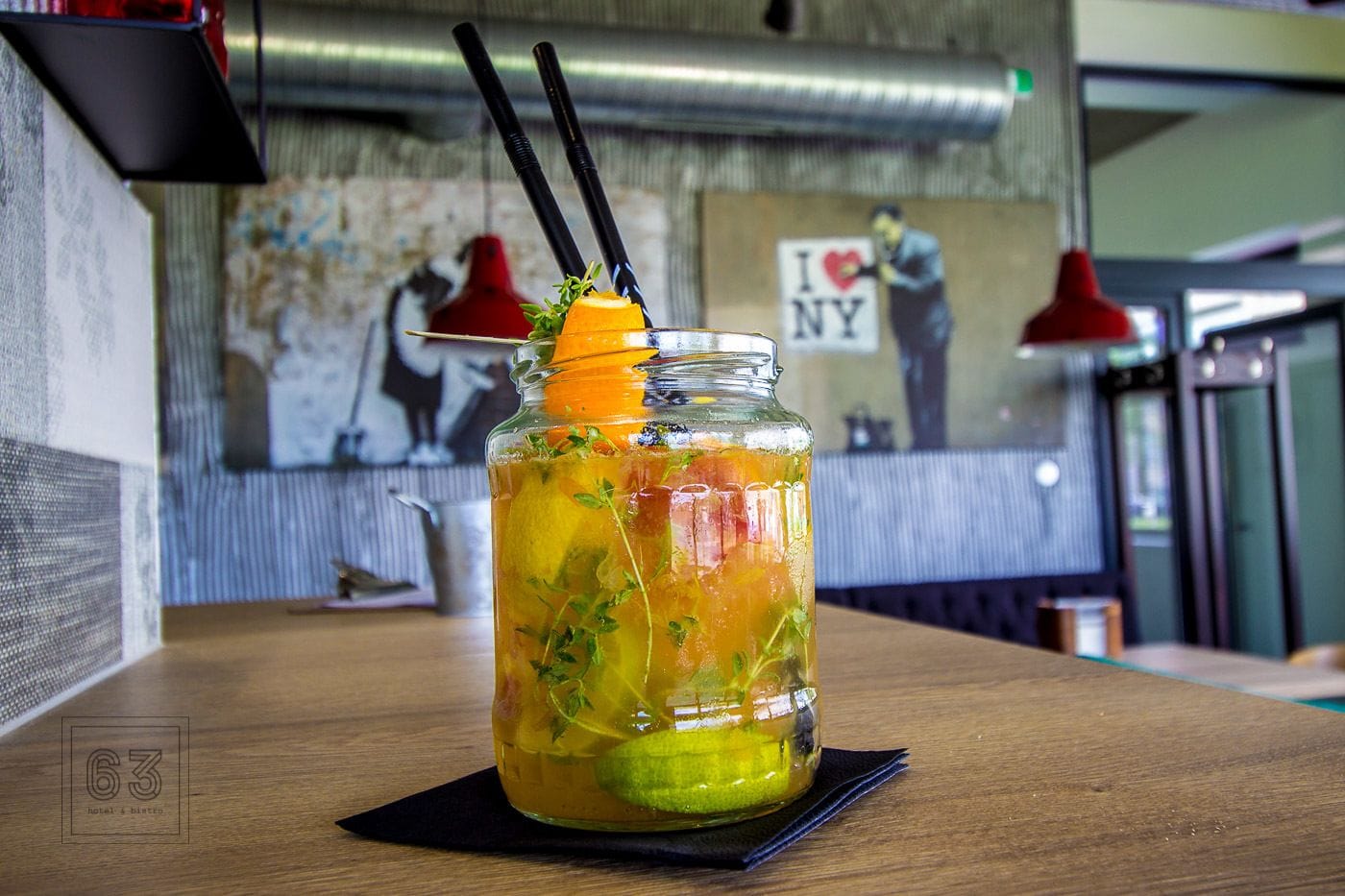 A glass jar with a drink garnished with herbs and citrus, placed on a wooden table in a restaurant setting.