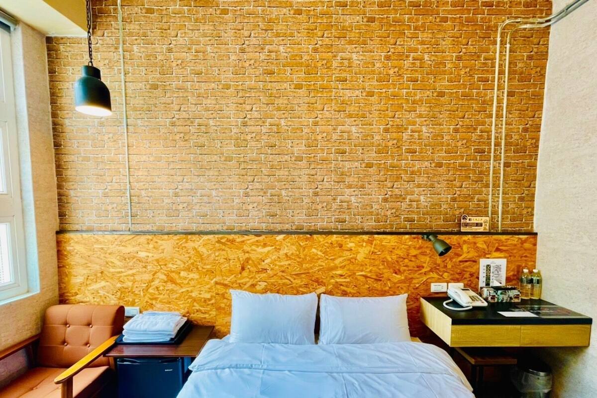 A hotel room with a brick wall, a bed with white bedding, a desk with a lamp, a chair, and a small trash bin.