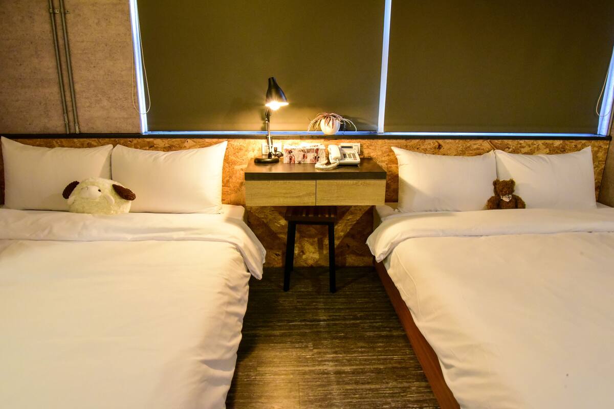 A hotel room with two beds, a desk with a lamp, a teddy bear, and a small table.