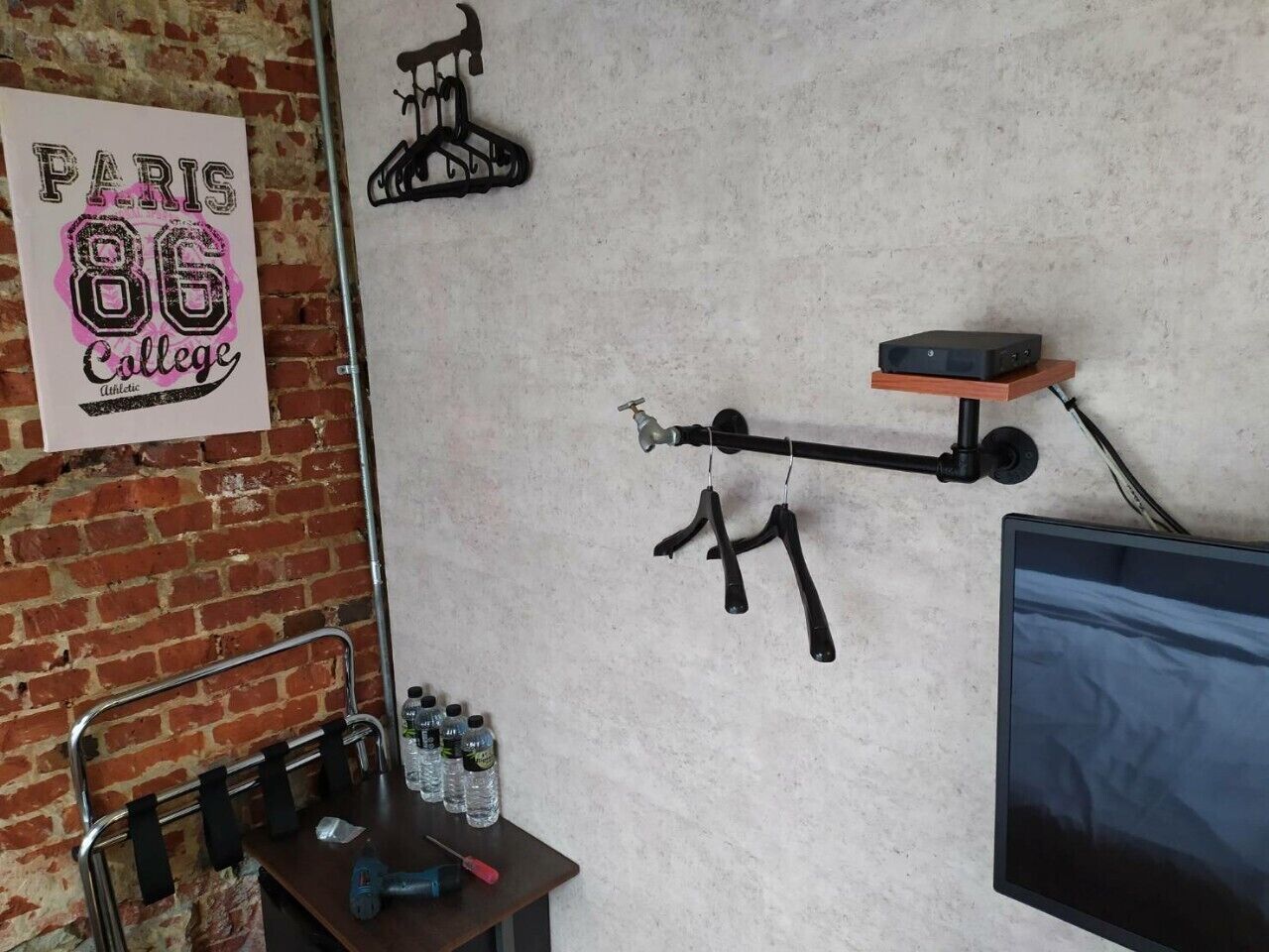 A room with a brick wall, a poster of Paris 86 College, a bicycle rack, a shelf with a TV, and a table with bottles and a power tool.