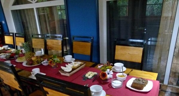 Un comedor con una larga mesa dispuesta para el desayuno, con platos de comida, tazas y un jarrón con flores.