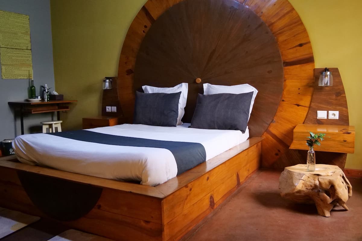 Una cama de madera con cabecera curvada, una mesita de noche y un taburete de madera con una planta.