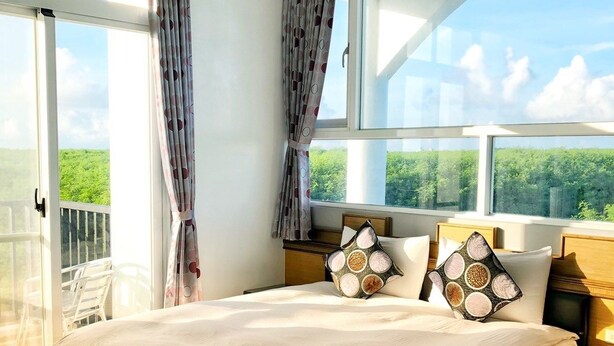 A spacious bedroom with a large bed, two windows with curtains, a balcony, and a view of greenery. - Moon Fish (Magong)