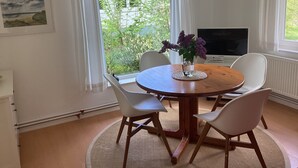 A dining area with a round wooden table, white chairs, a vase with purple flowers, a television, and a view of greenery outside. - Crow's Nest - Bungalow 2 - Ferienhäuser Pönitz am See (Scharbeutz)
