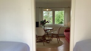 A room with two beds, a dining table with chairs, a red sofa, and a window with a view of greenery. - Crow's Nest - Bungalow 2 - Ferienhäuser Pönitz am See (Scharbeutz)