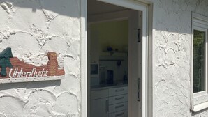 A white building with a wooden door and a window. Inside, there is a kitchen with cabinets and a countertop. - Crow's Nest - Bungalow 2 - Ferienhäuser Pönitz am See (Scharbeutz)