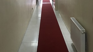Un long couloir étroit, doté d’une moquette rouge, de carreaux blancs au sol et de murs ornés de tableaux encadrés.