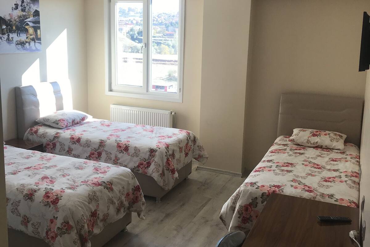 A bedroom with two beds, a wooden desk, a radiator, and a window with a view of the outside.