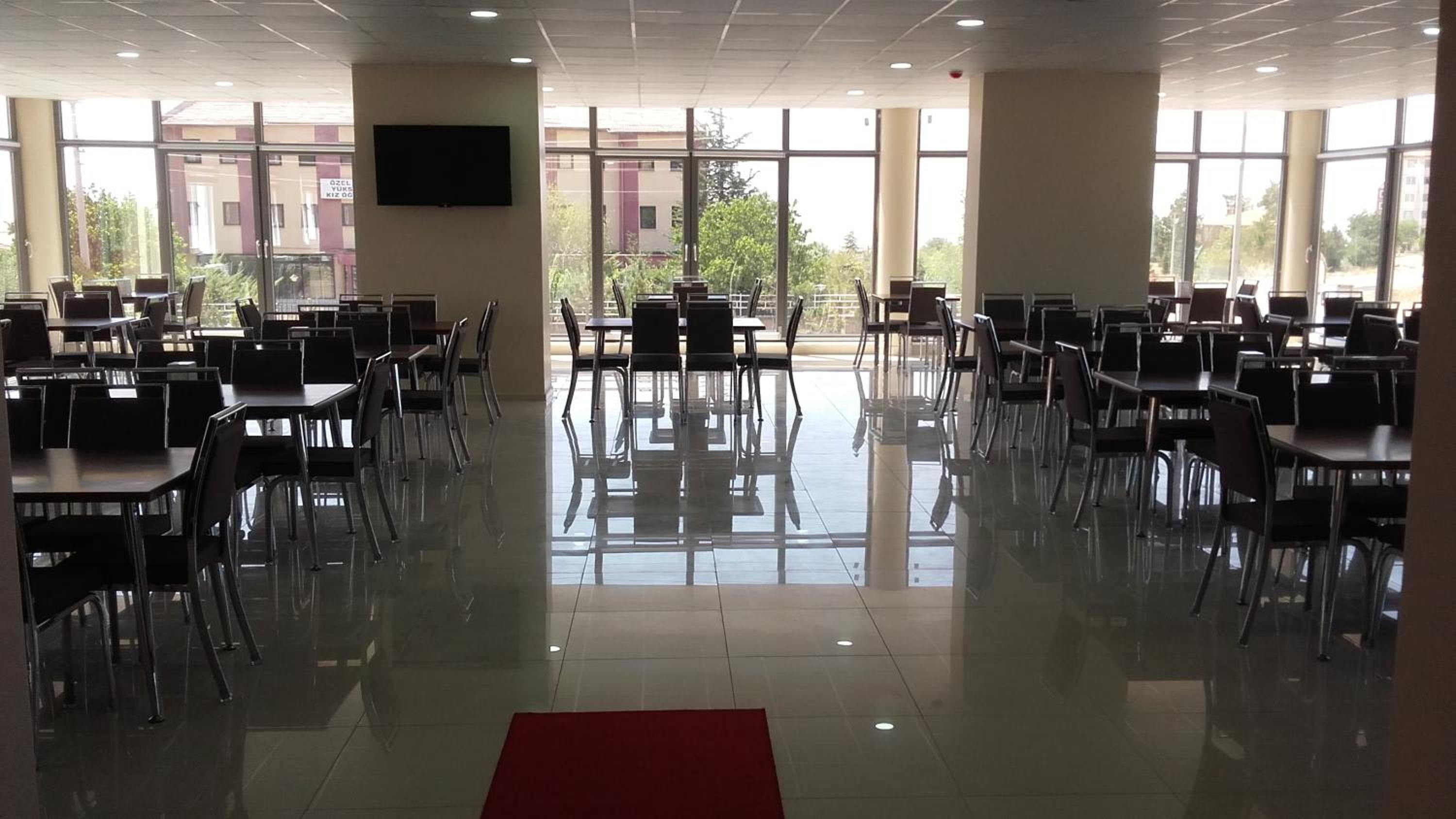 A spacious indoor area with multiple tables and chairs arranged in rows, large windows allowing natural light, and a flat-screen TV mounted on the wall.