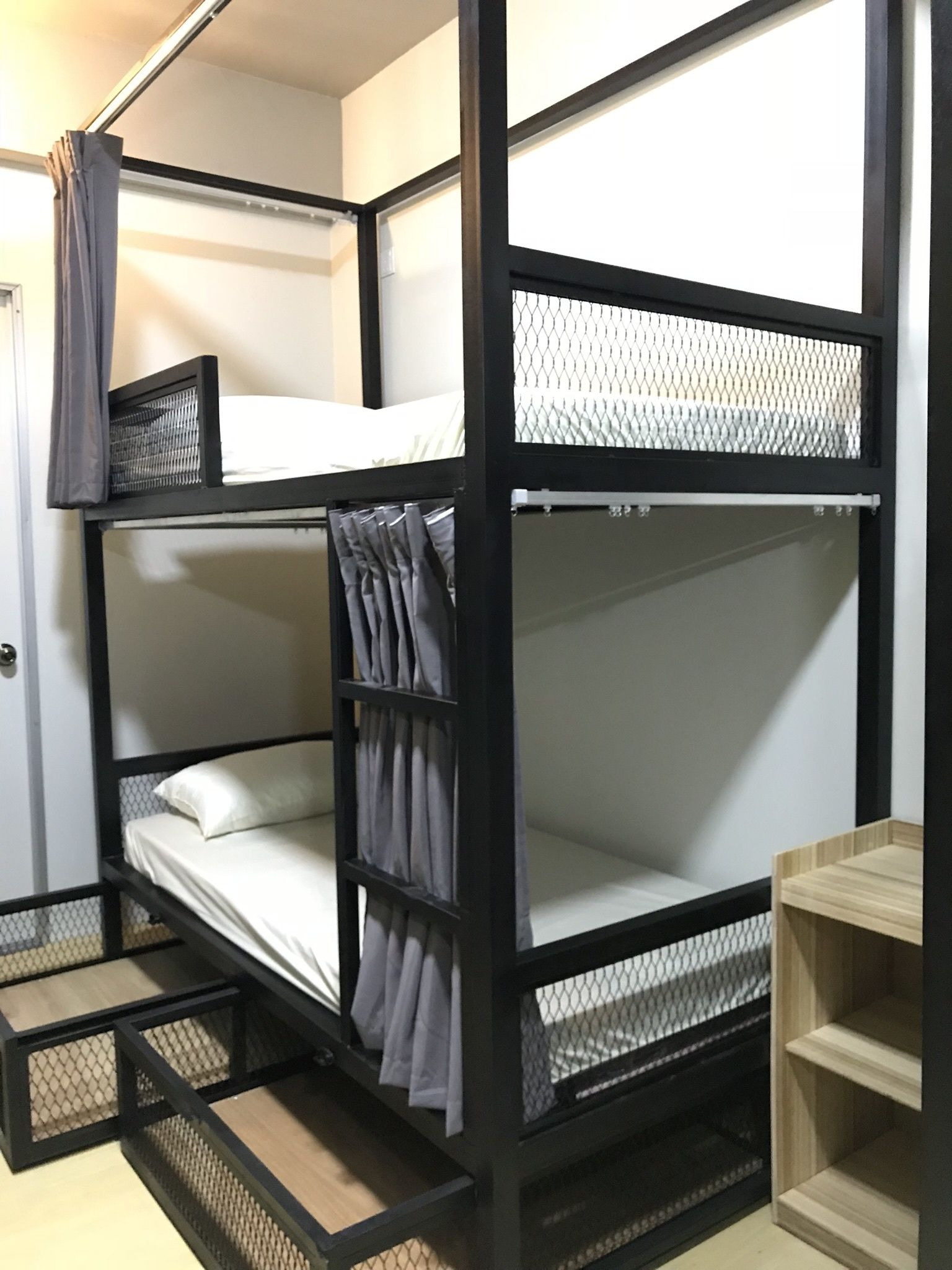 Una cama litera con una cama inferior y una superior, ambas con laterales de malla. Hay un pequeño estante de madera en el lado derecho de la cama.