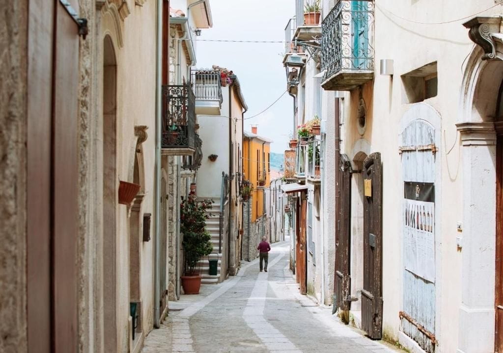 A narrow street lined with buildings, a person walking down the street, and balconies with plants.