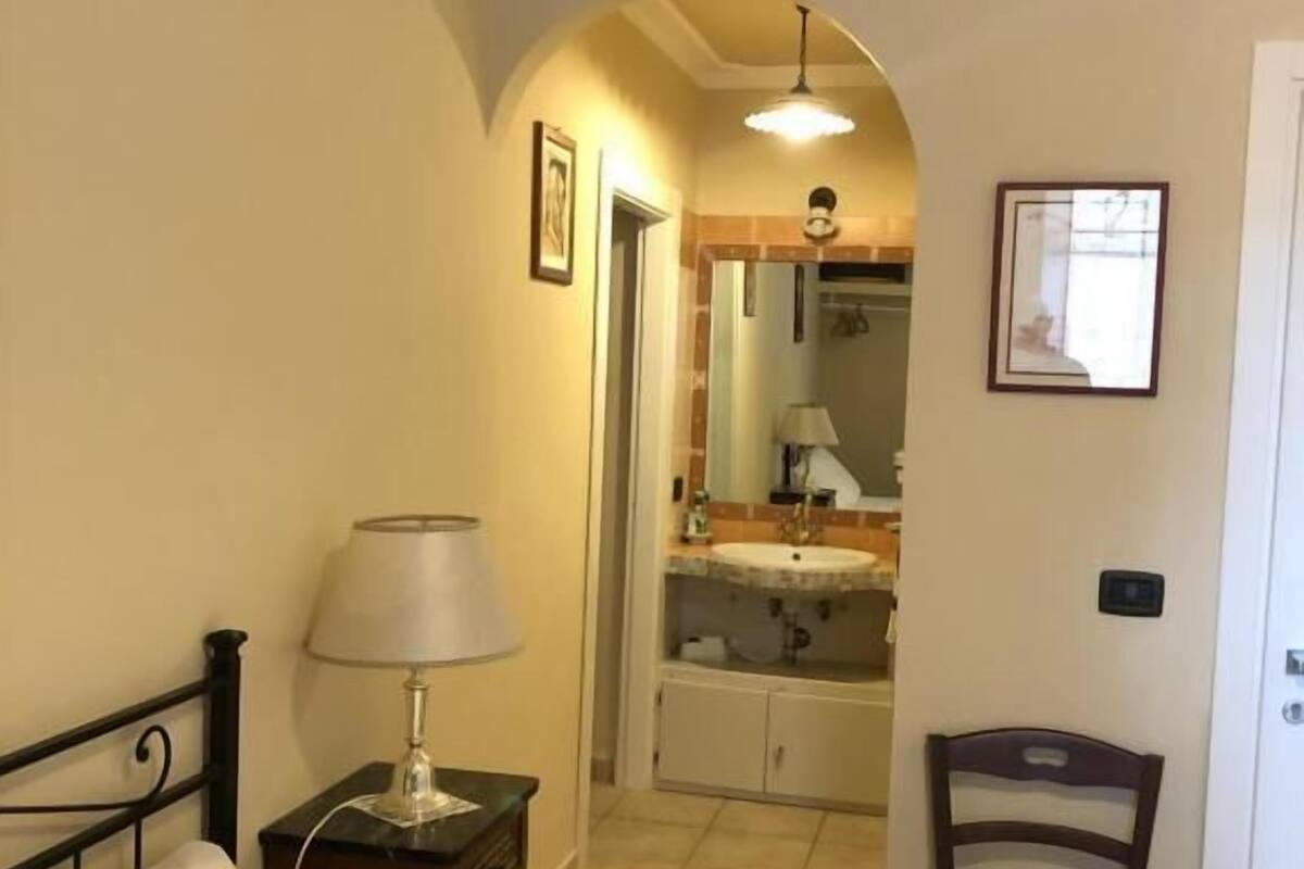 A bedroom with a bed, a nightstand with a lamp, a chair, and a bathroom visible through an archway.