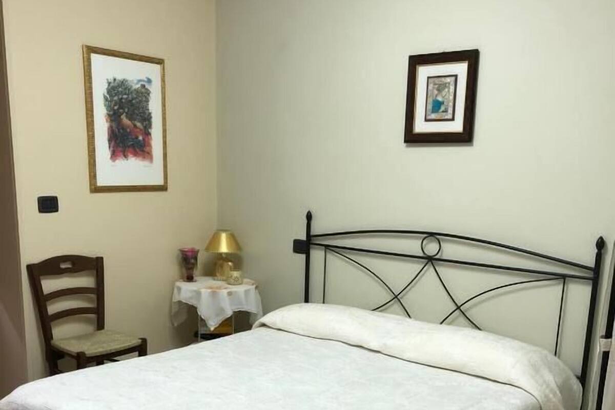 A neatly made bed with a white bedspread, a small table with a lamp, and two framed pictures on the wall.