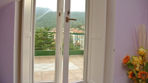 Una stanza con una porta scorrevole in vetro che dà su un balcone con vista su un paesaggio verde e degli edifici.