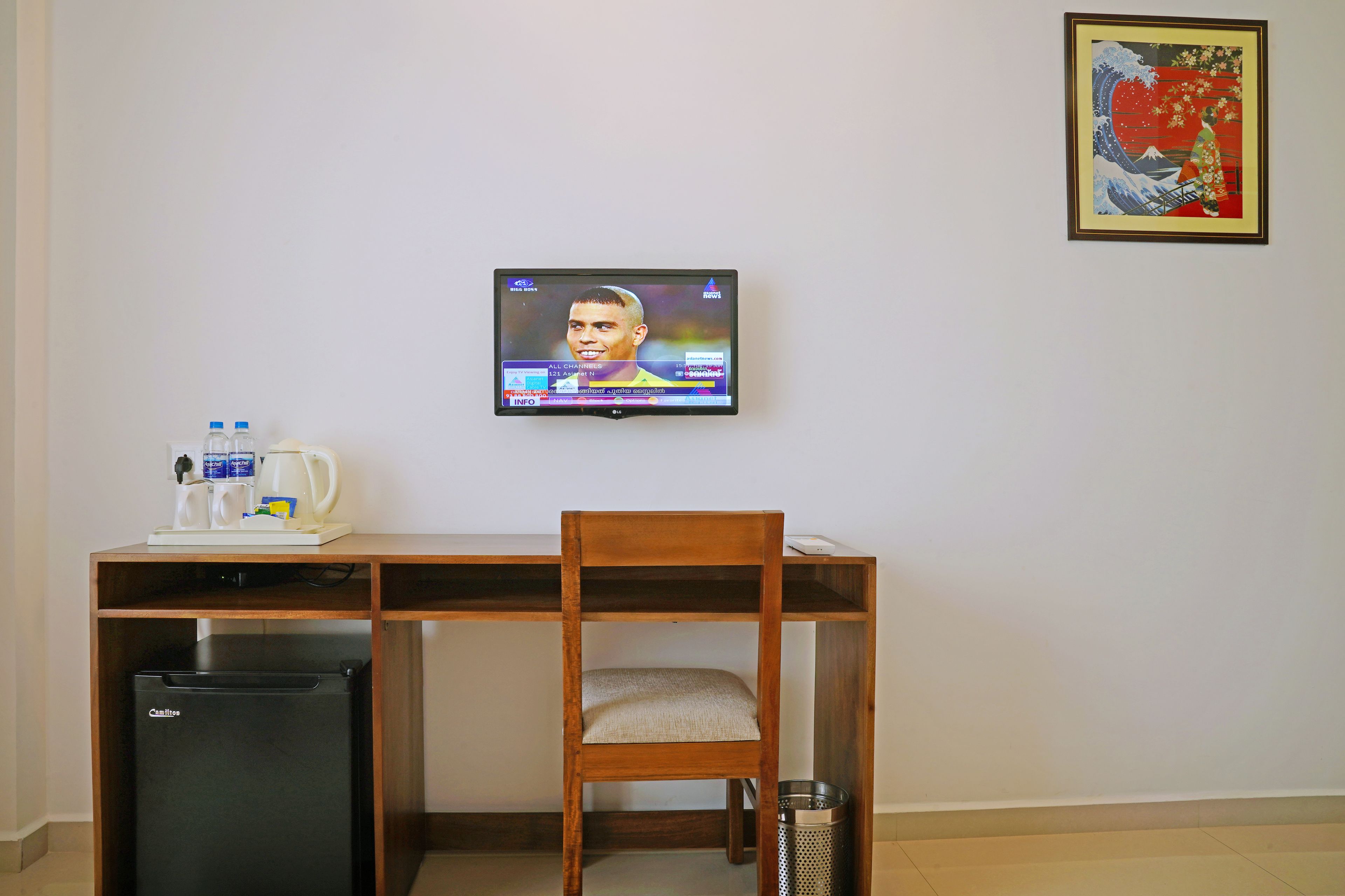 A room with a wooden desk, a black refrigerator, a chair, a TV mounted on the wall, and a trash can.
