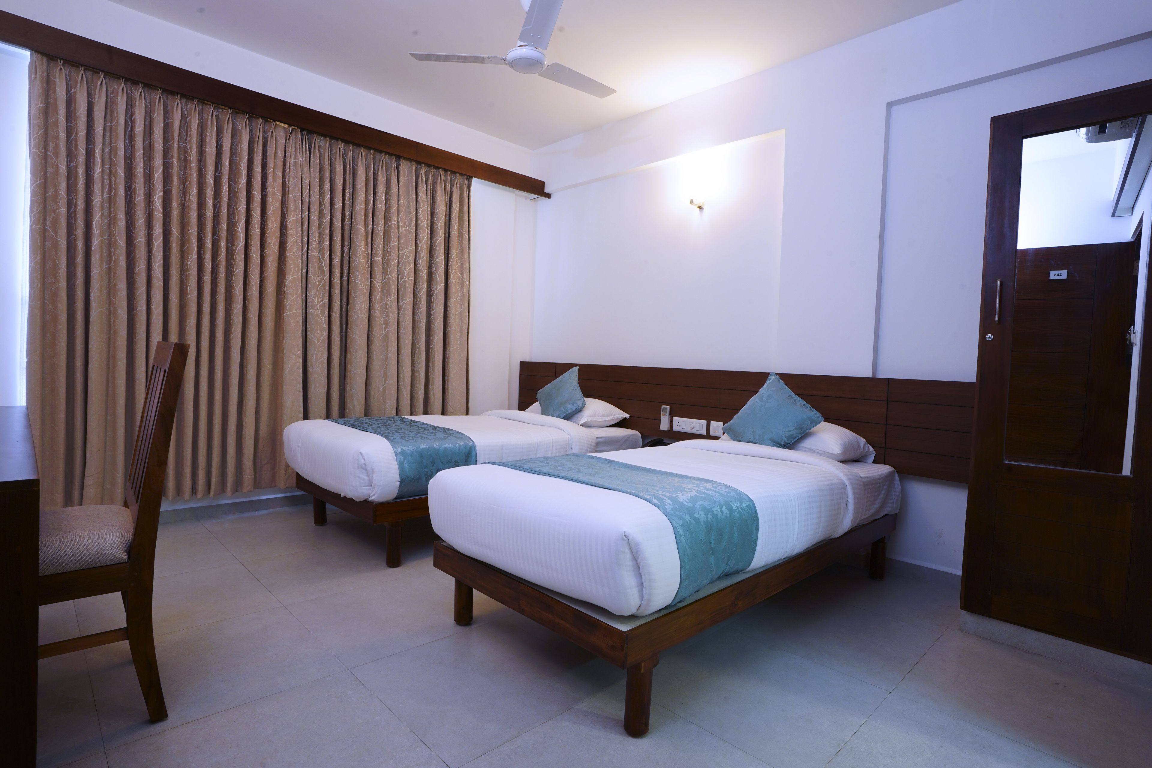 A hotel room with two beds, a ceiling fan, a wooden chair, and a large window with curtains.