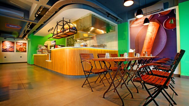 A restaurant with a curved counter, wooden chairs, and a green wall with a large food advertisement. - Niko Hotels (Kanayannur)