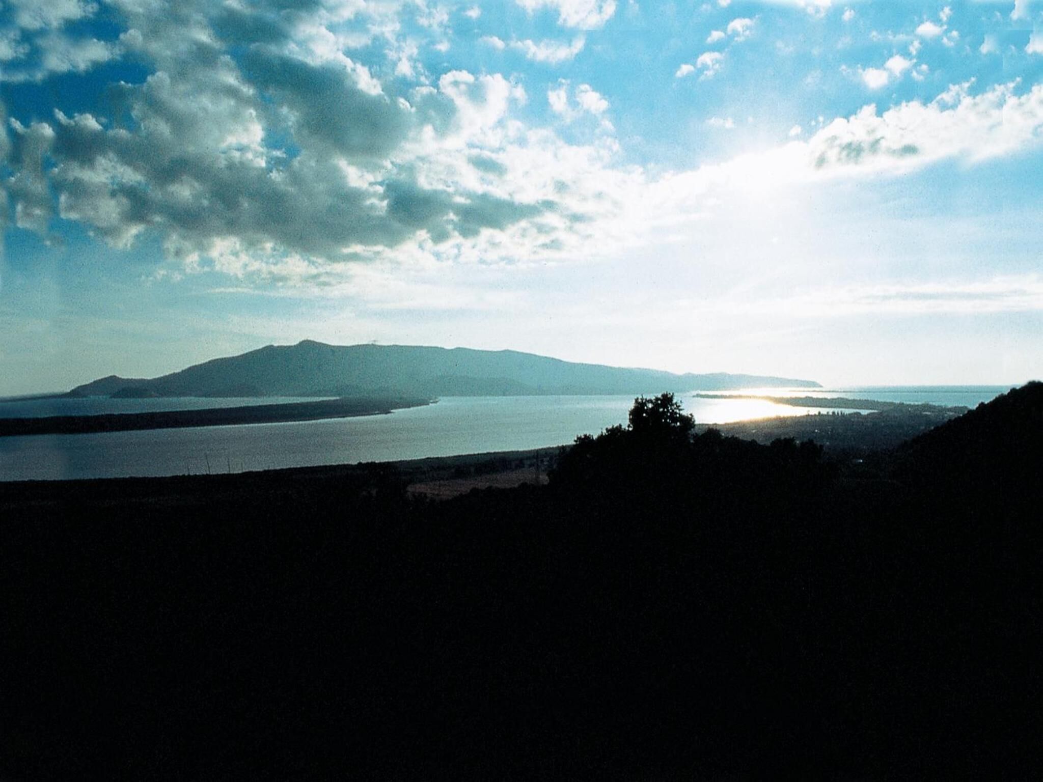 Vista panoramica di un paesaggio costiero con un corpo d'acqua, montagne in lontananza e cielo sereno.