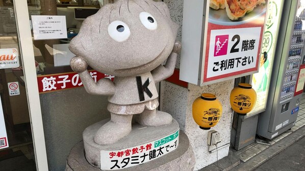 Una estatua de piedra de un personaje con la letra ‘K’ en el pecho, ubicada sobre un pedestal con un cartel que indica que es una estatua de un personaje llamado ‘Satan’. 