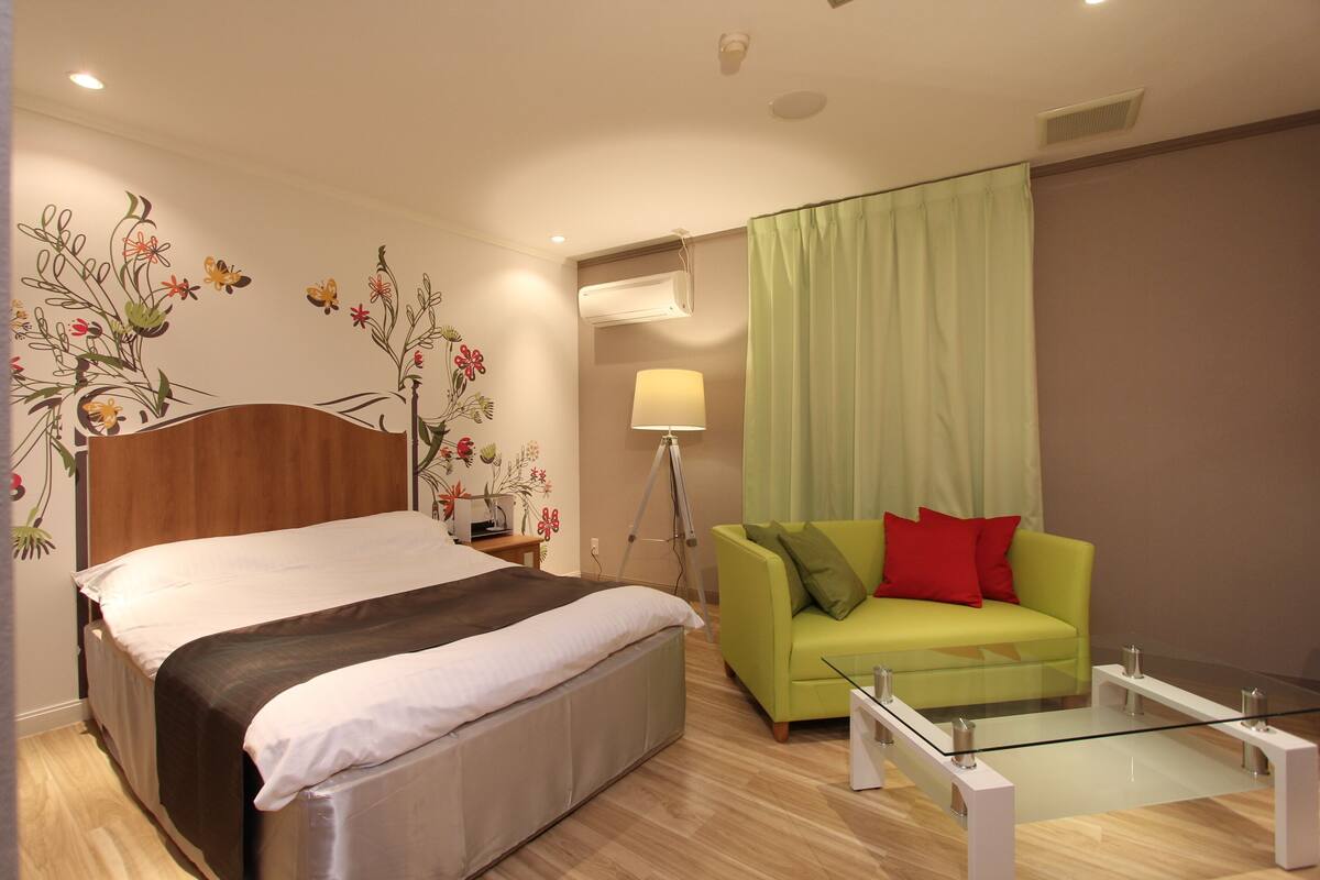 Une chambre d’hôtel moderne avec un lit, un canapé vert, une table basse en verre et un mur décoré d’œuvres d’art florales.