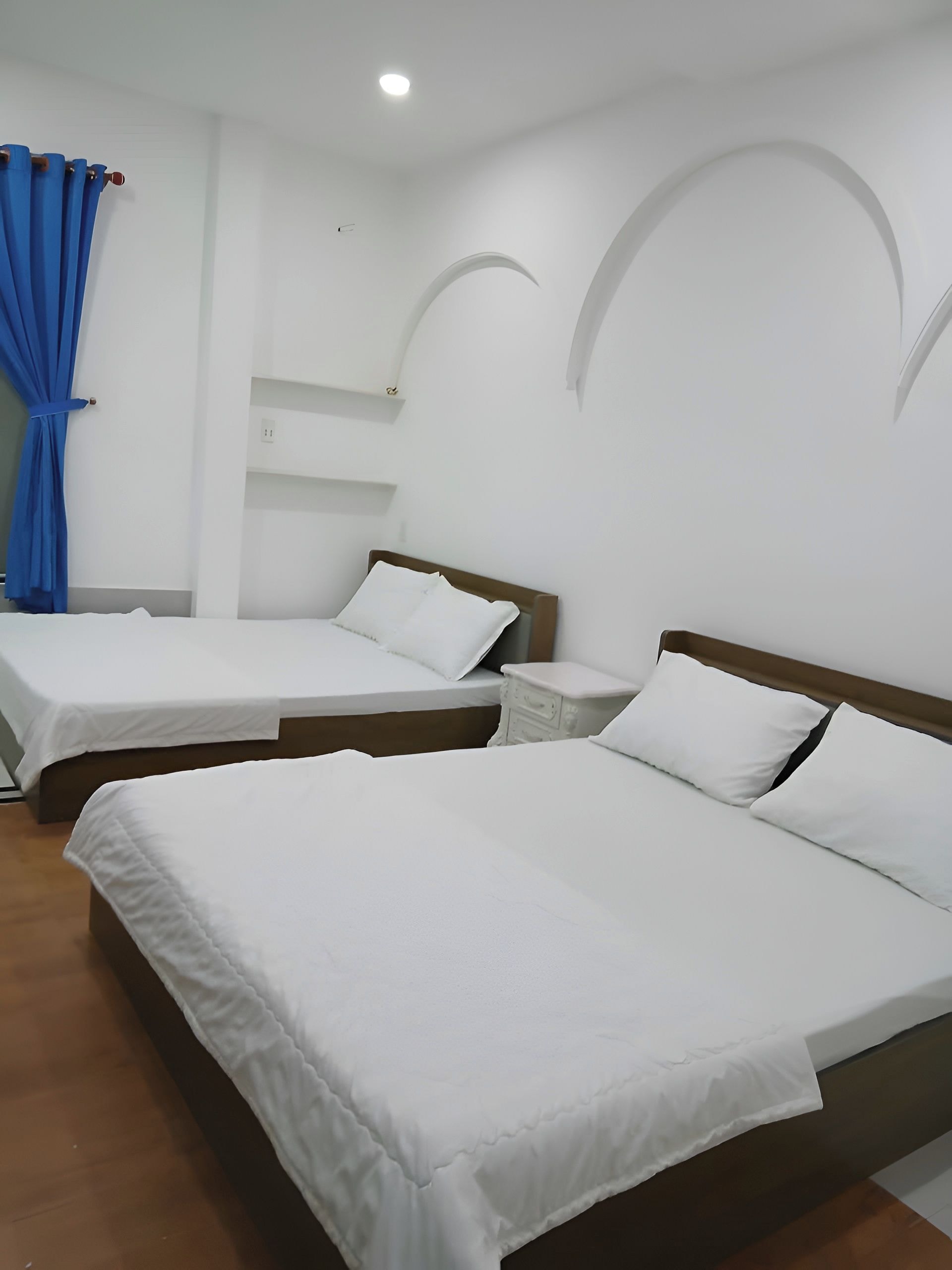 Habitació d'hotel amb dos llits, una tauleta de nit, una cortina blava i un sostre blanc amb il·luminació empotrada.