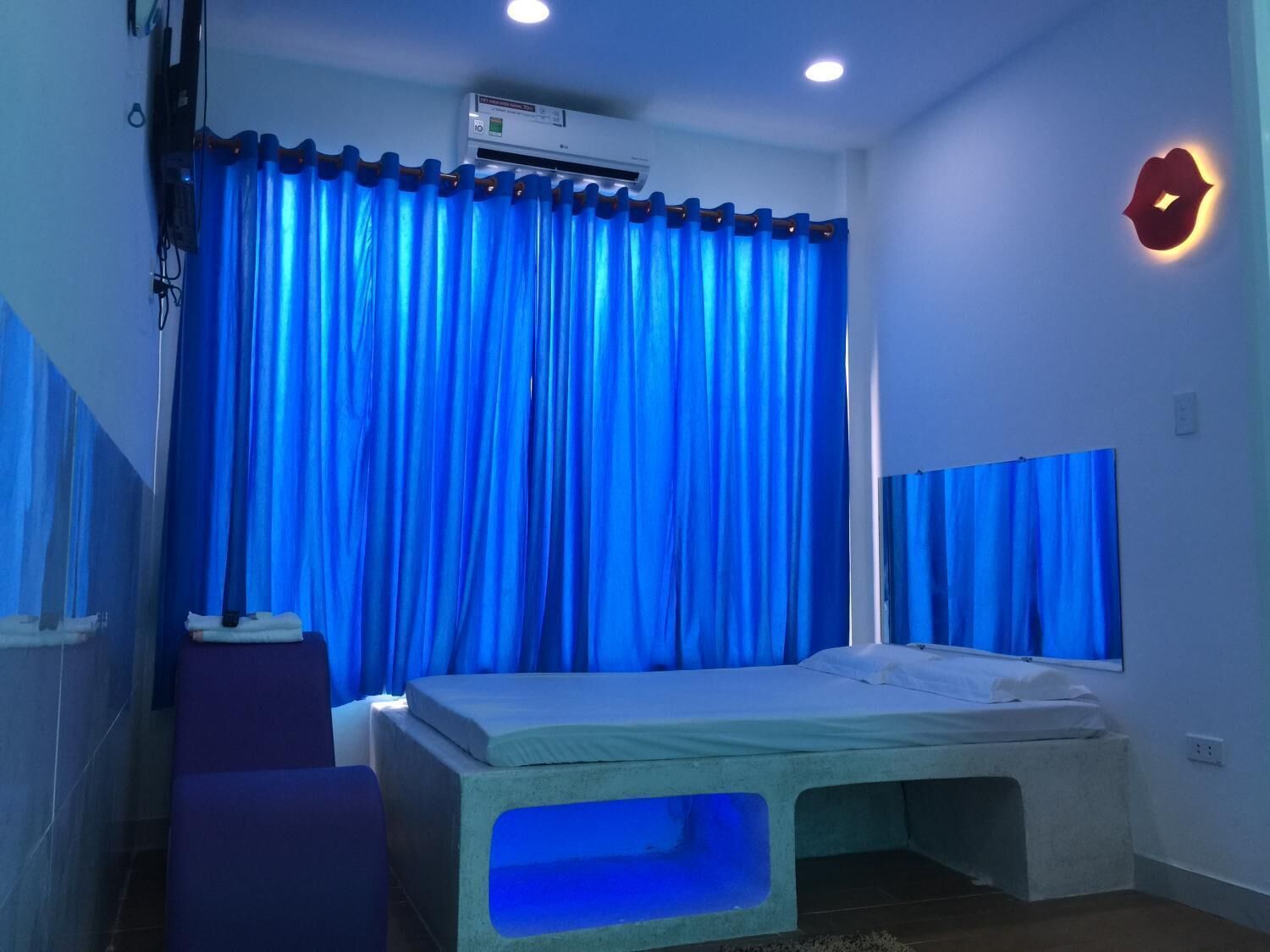 A room with a bed, a window with blue curtains, a wall-mounted air conditioner, and a small table with a cushion.