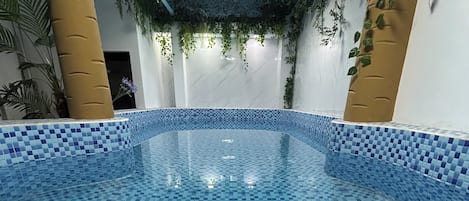 An indoor swimming pool with blue mosaic tiles, surrounded by walls with vertical greenery and a ceiling with hanging plants.