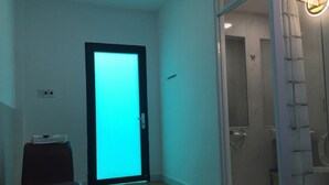 Hotelkamer met een blauwgroene deur, een bed, een klein tafeltje met een stapel papieren, en een badkamer met douche en toilet.