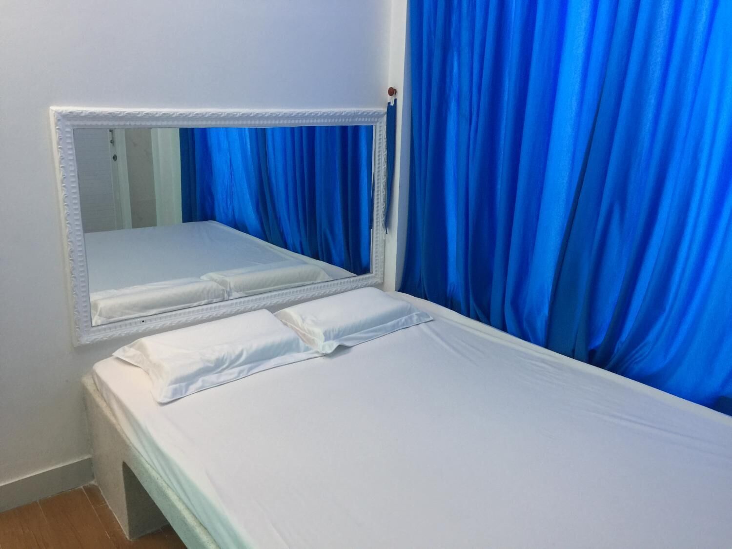 A single bed with white bedding, a large mirror with a white frame, and blue curtains.