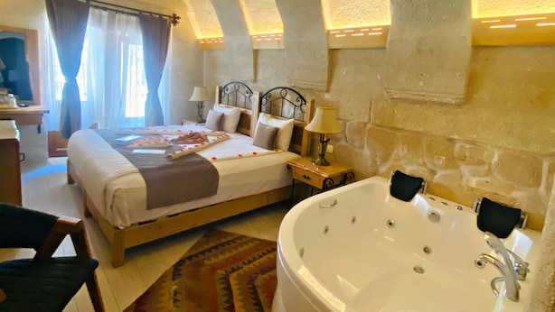 A bedroom with a large bathtub, a bed with pillows, a nightstand, and a window with curtains.