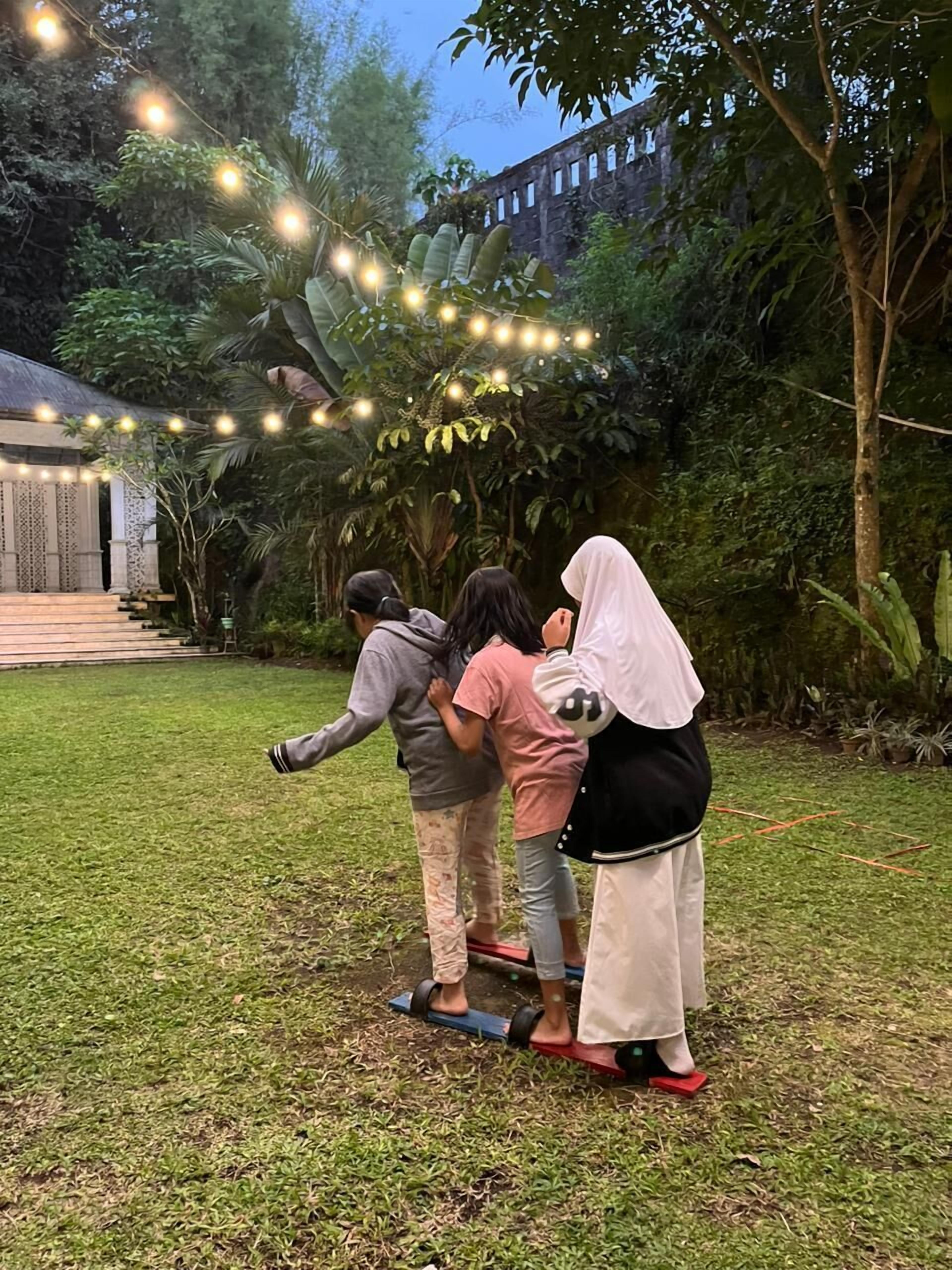 A group of people playing on a seesaw in a garden with string lights overhead.