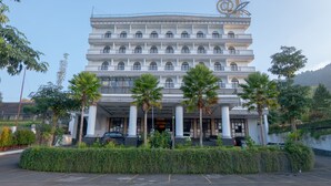 Un hotel de varios pisos con entrada central, palmeras y un estacionamiento.