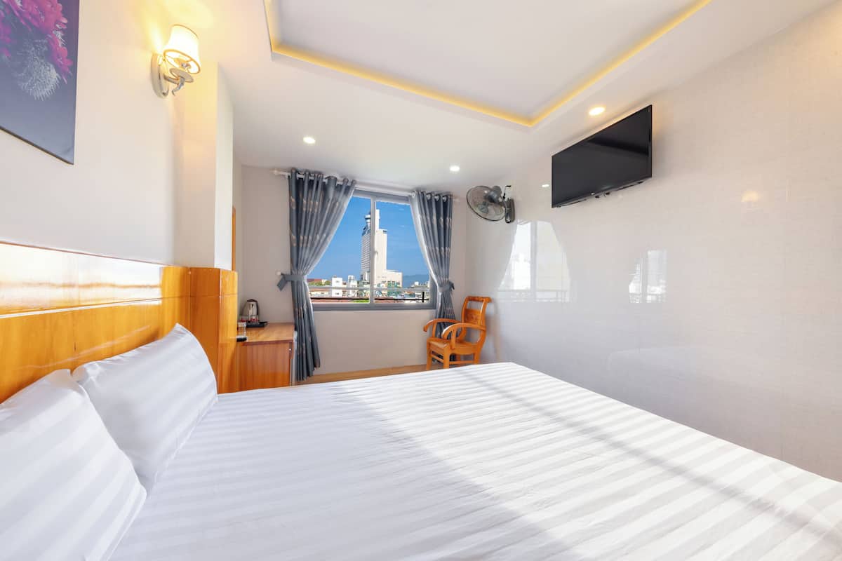 Une chambre d’hôtel avec un lit, un téléviseur fixé au mur, une fenêtre avec des rideaux et une vue sur un bâtiment à l’extérieur.