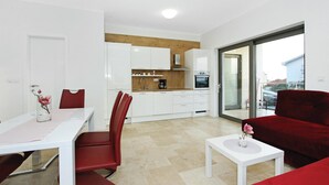 Une cuisine moderne avec des armoires blanches, un plan de travail en bois et un four encastré. On y trouve un coin repas avec une table blanche et des chaises rouges. Un canapé rouge est visible au premier plan.