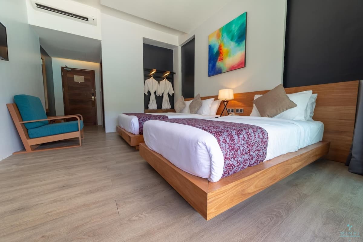 A hotel room with two beds, a sitting area with a chair, a painting on the wall, and a wooden floor.