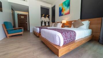 Habitación de hotel con dos camas, un rincón de descanso con una silla, un cuadro en la pared y suelo de madera.
