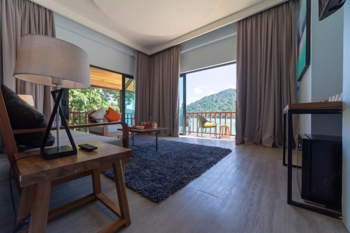 A modern hotel room with a wooden desk, a lamp, a TV, and a balcony with a view of greenery and hills.