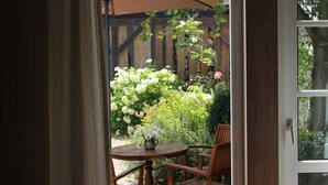 Une porte ouverte donne sur un patio aménagé avec une table et des chaises, entouré de verdure et doté d’un sol en briques.