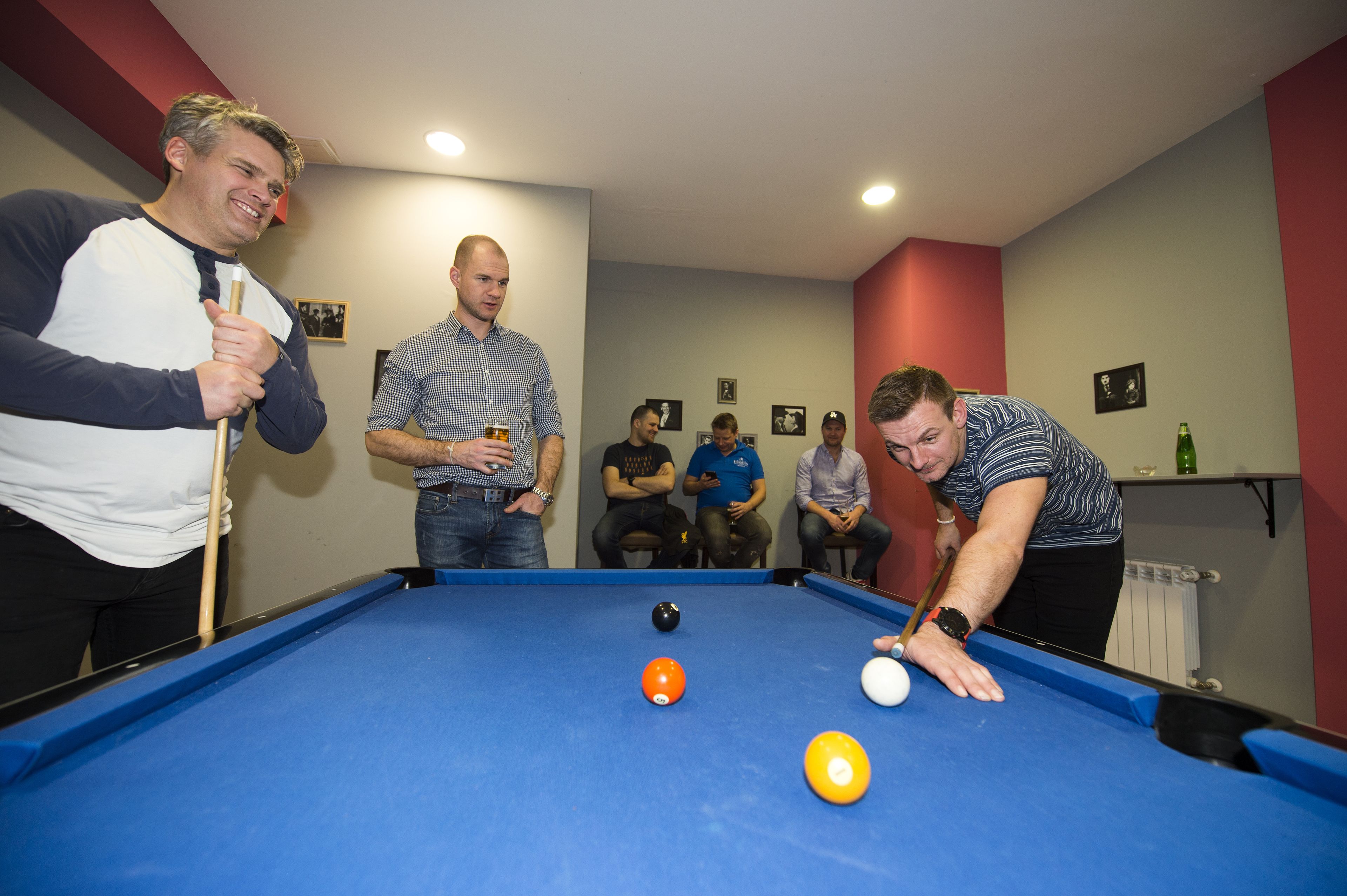 A pool table in a room with a red accent wall and a shelf with bottles.